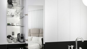 Een moderne hotelkamer met een kleurenspel van wit en zwart, met een grote spiegel, een bed met een witte hoofdbord, een witte wastafel met een moderne kraan, en een zwarte stoel.