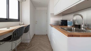 Een kleine keuken met een houten aanrecht, een roestvrijstalen spoelbak en een zwarte waterkoker. Er staan twee stoelen en er is een deur naar een andere kamer.