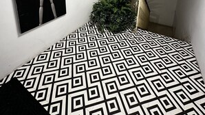 Een kamer met een vloer met een geometrisch patroon, een plafondlamp, een speaker bevestigd aan de muur, een grote potplant en een ingelijst beeld.