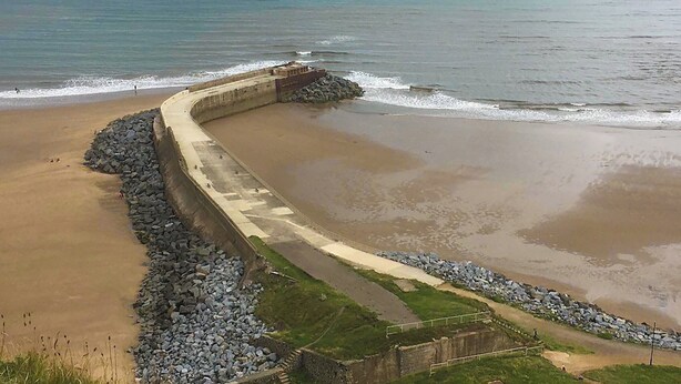 Une digue marine en béton incurvée, dotée d’une base rocheuse, qui mène à une plage de sable.