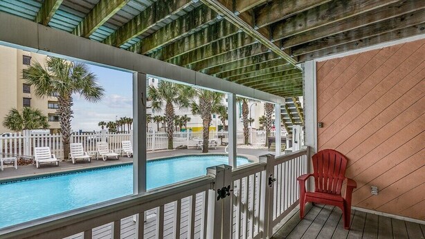 A poolside area with a wooden deck, a red chair, and a view of a building with palm trees.