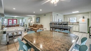 A spacious living area with a pool table, a dining table with chairs, a ceiling fan, and a kitchen area with white cabinets and a refrigerator.