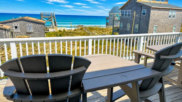 Een houten terras met een tafel en stoelen, met uitzicht op een strand en gebouwen in de verte.