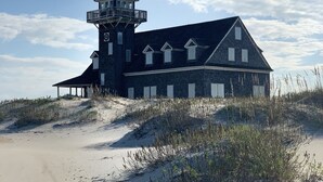 Een groot, donker gebouw met een toren en meerdere ramen, gelegen aan een zandstrand met weinig beplanting.
