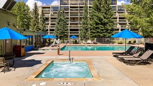 Área de piscina com espreguiçadeiras e guarda-sóis, cercada por árvores altas e um edifício de vários andares ao fundo.