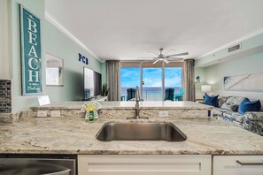 A modern kitchen with a large sink, a ceiling fan, and a view of the ocean through large windows. - Tidewater 713- FREE Beach Service for 4, Golf+Perks, 2BR/3BA+Bunks,Pools,Gym! (Panama City Beach)