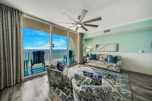 A spacious living room with a large sliding glass door leading to a balcony with a view of the ocean. There is a ceiling fan, a patterned sofa set, and a coffee table. - Tidewater 713- FREE Beach Service for 4, Golf+Perks, 2BR/3BA+Bunks,Pools,Gym! (Panama City Beach)