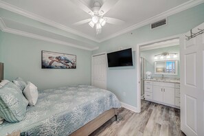 A bedroom with a bed, a ceiling fan, a TV, a bathroom with a sink and mirror, and a hallway. - Tidewater 713- FREE Beach Service for 4, Golf+Perks, 2BR/3BA+Bunks,Pools,Gym! (Panama City Beach)