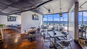 Um restaurante com mesas, cadeiras, janelas amplas e vista para o mar.