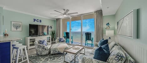 A living room with a TV, a ceiling fan, a sofa, a coffee table, and a view of the ocean through large windows.