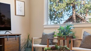 Un salon avec un canapé, une table basse agrémentée de plantes, un téléviseur posé sur un meuble et une fenêtre donnant sur des arbres.