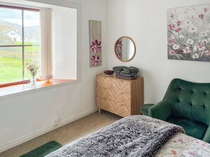 Quarto com uma janela grande, uma poltrona estofada verde, cômoda de madeira e um quadro com flores.