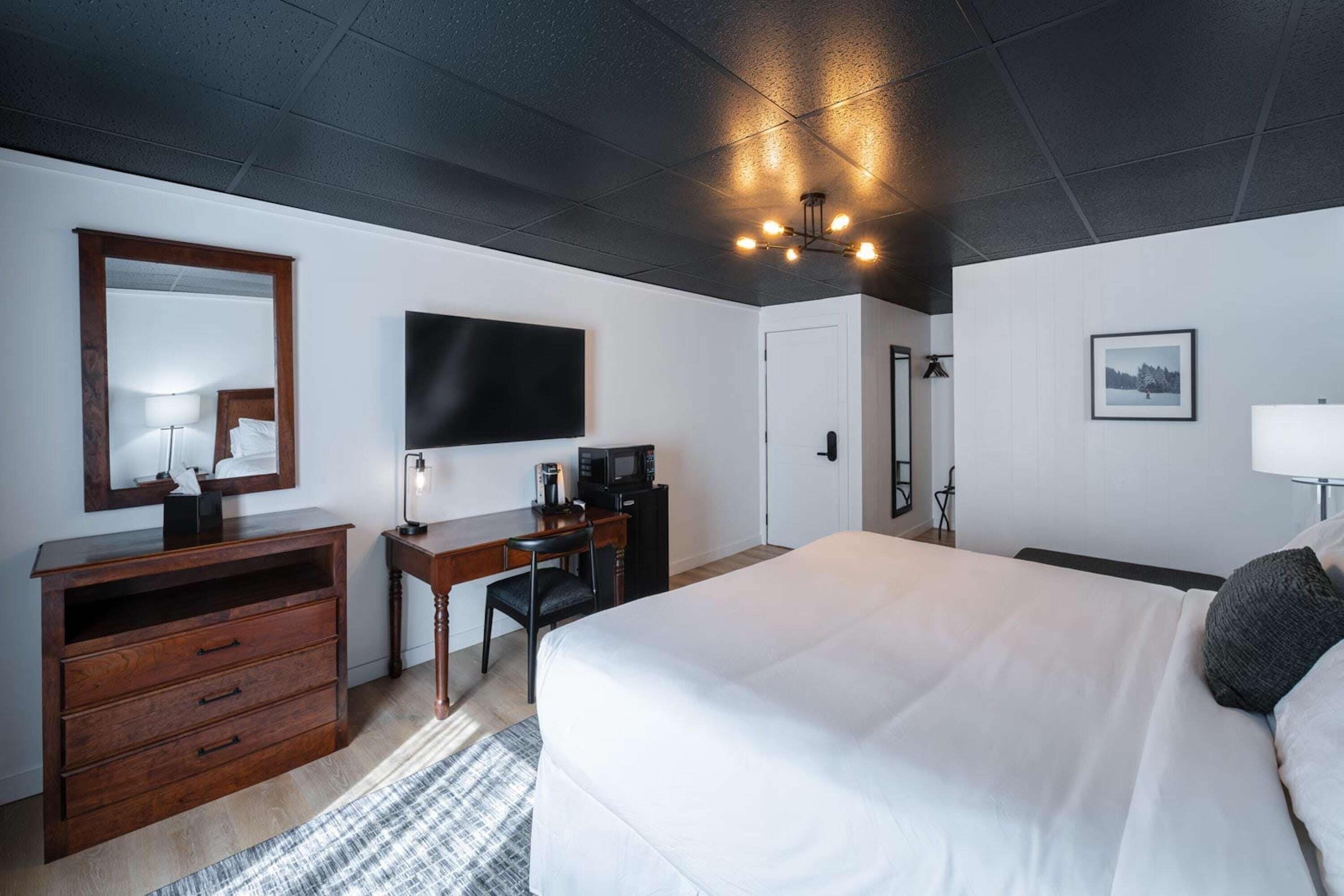 A hotel room with a large bed, a desk with a chair, a flat-screen TV, a dresser, and a ceiling light fixture.