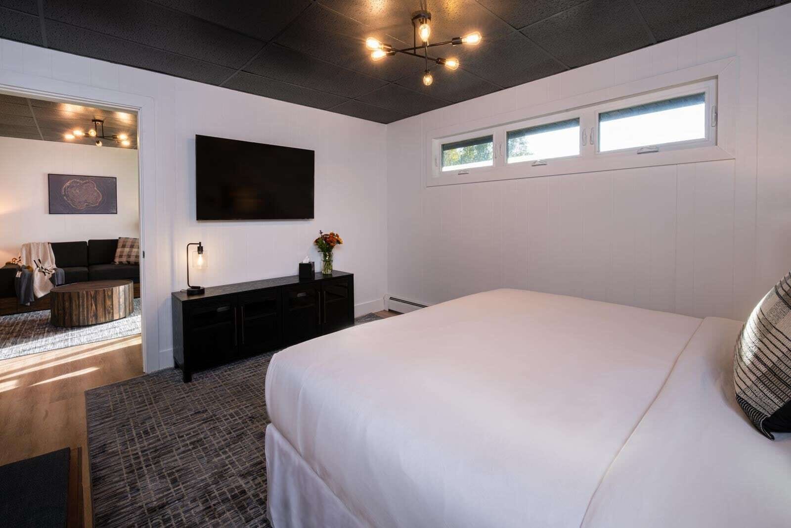 A modern bedroom with a large bed, a flat-screen TV mounted on the wall, a small bedside table with a lamp, and a window with two small panes.