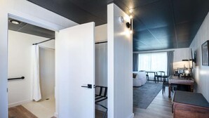 A modern hotel room with a wooden floor, a white door leading to a bathroom, a seating area with a sofa and a table, and a desk with a lamp. - Trailhead by Weekender (Tupper Lake)