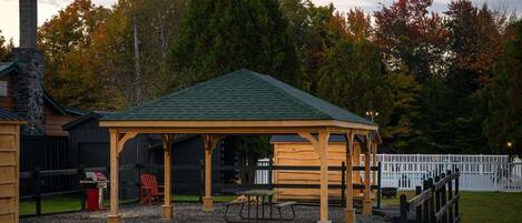 Un kiosque en bois avec un toit vert, une table de pique-nique et un barbecue, le tout situé dans un parc.