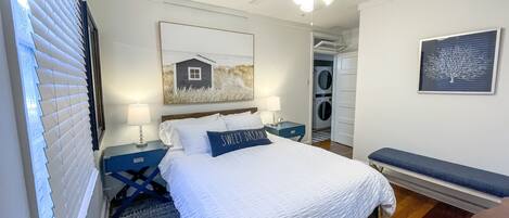 Une chambre à coucher comprenant un lit, des tables de chevet, un ventilateur de plafond, une fenêtre avec des stores, une œuvre d’art accrochée au mur et une porte donnant sur une buanderie.