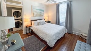 A hotel room with a bed, a ceiling fan, a window with curtains, a wall-mounted painting, and a washing machine in the corner.