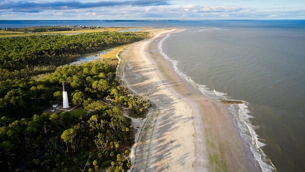 An aerial view of a coastal area with a sandy beach, dense green forest, and a lighthouse.