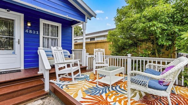 A blue house with a porch, white wicker furniture, and a colorful patio rug.