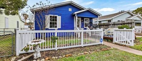 A single-story house with a blue exterior, a white picket fence, and a small garden area with a birdbath.