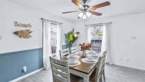 A dining area with a wooden table set for four, white chairs, a ceiling fan, and a wall-mounted decorative piece.