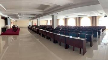 A conference room with rows of tables and chairs, a stage with a red carpet, and large windows with curtains.