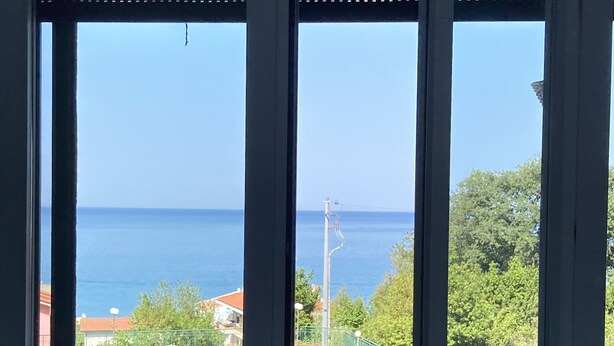 Ein Balkon mit Korbmöbeln, einem roten Stuhl und Blick auf das Meer und viel Grün.