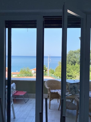 Un balcon agrémenté de meubles en osier, d’un fauteuil rouge et offrant une vue sur la mer et la verdure.