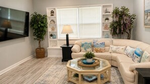 A living room with a beige sofa, a glass coffee table, a TV, and a bookshelf with decorative items. - Cozy home in quiet neighborhood!! LLC.              2 Kings  bed and 1 Queen  (Spring Hill)