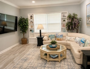 A living room with a beige sofa, a glass coffee table, a TV, and a bookshelf with decorative items.
