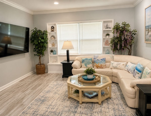 A living room with a beige sofa, a glass coffee table, a TV, and a bookshelf with decorative items.