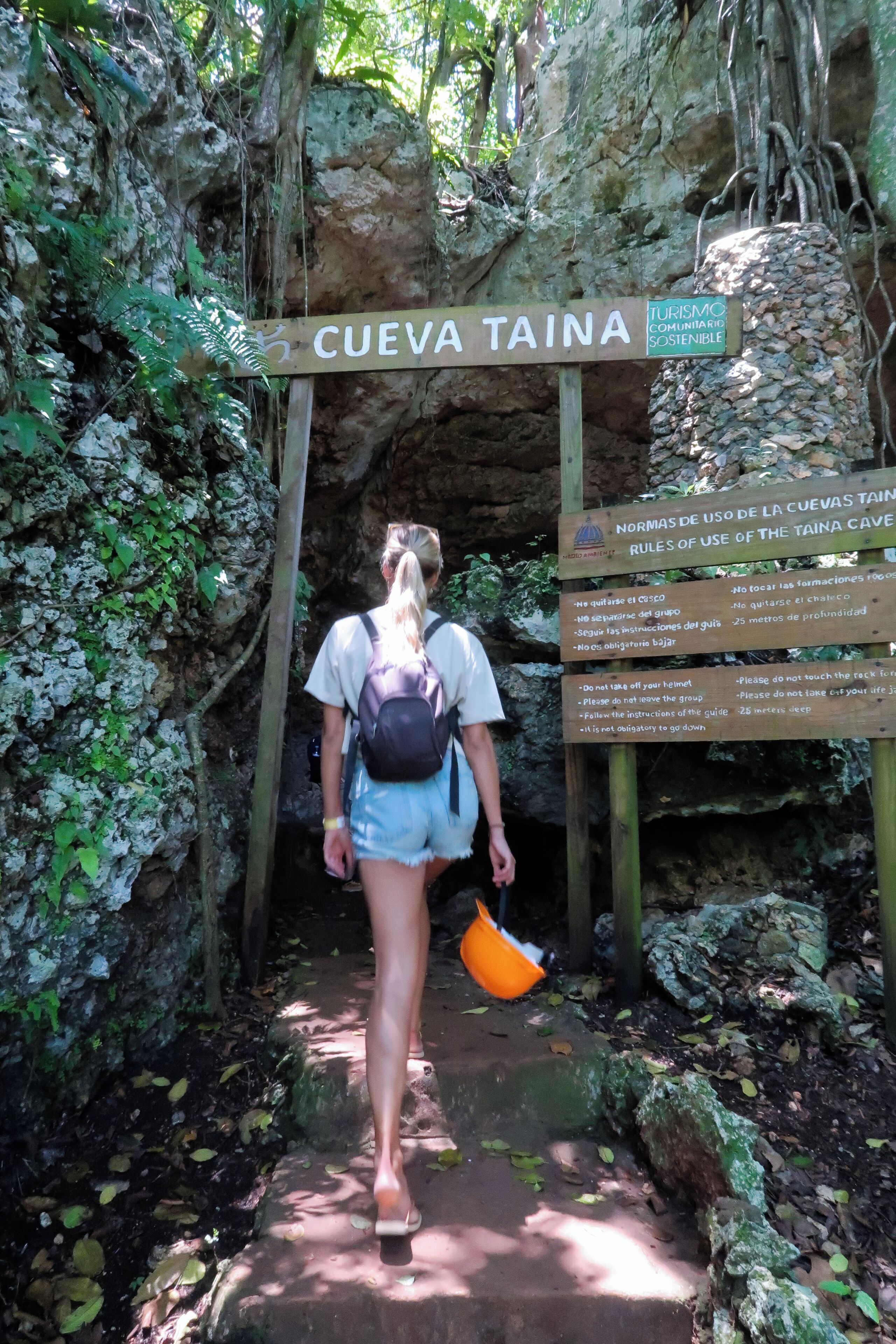A person walking towards a cave entrance labeled 'CUEVA TAINA' with a sign detailing usage rules.
