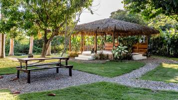 Un kiosque doté d’un toit de chaume, entouré d’un jardin bien entretenu avec un chemin de gravier, des bancs et diverses plantes.