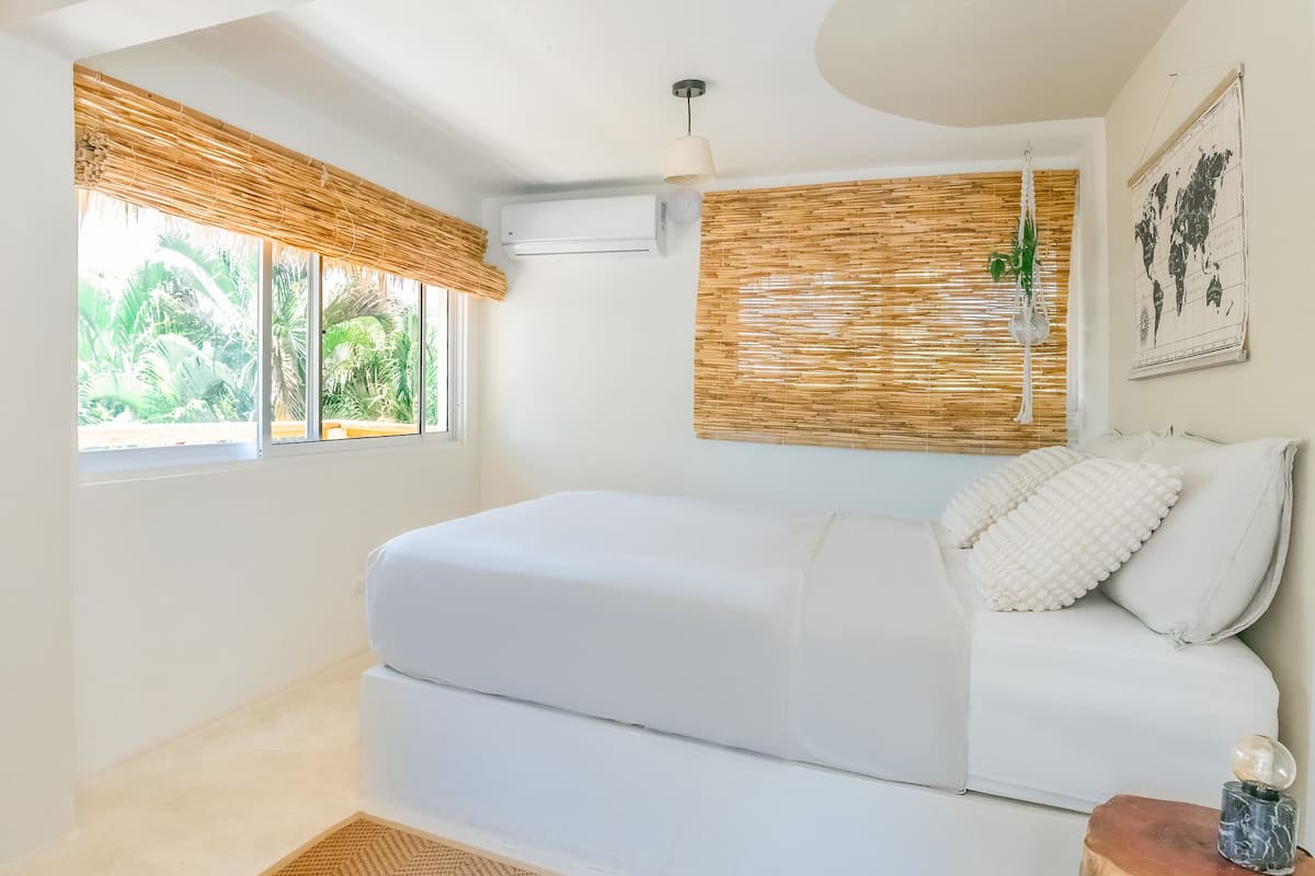 A bedroom with a bed, a window with a view of greenery, an air conditioning unit, and a world map on the wall.