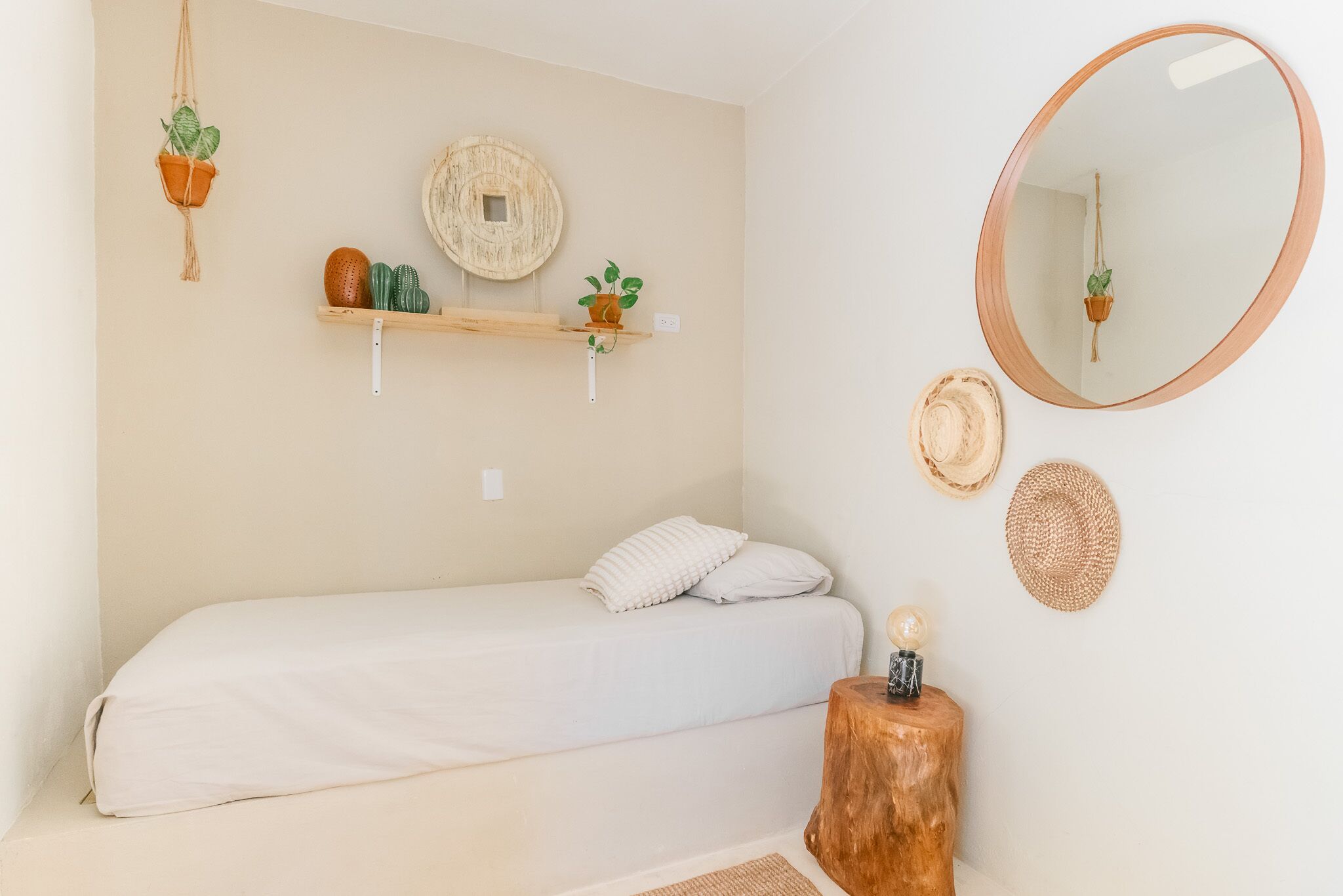 A bedroom with a single bed, a wooden stool with a plant, a round mirror, and wall-mounted shelves with plants and decorative items.