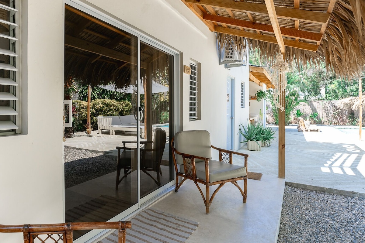 A covered outdoor seating area with a pool, a thatched roof, and a sliding glass door leading to a poolside lounge.