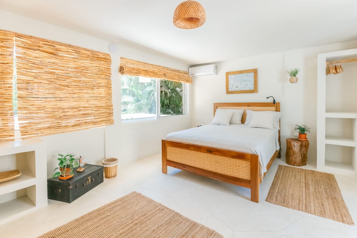 A modern bedroom with a wooden bed, a large window with bamboo blinds, a small side table with a plant, and a wall-mounted air conditioner.