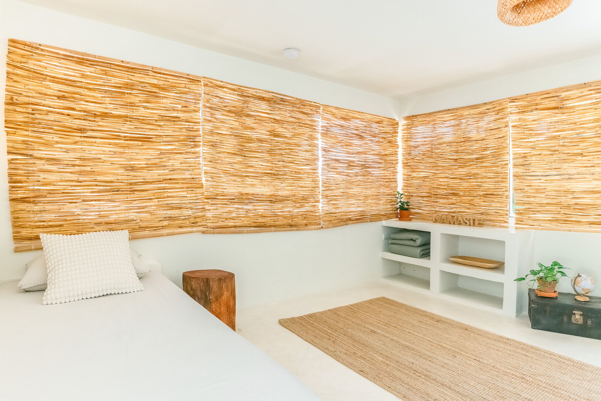 Un dormitorio con cama, mesita de noche de madera, cabecero tejido, estante con adornos y alfombra de fibras naturales.
