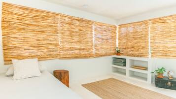 Un dormitorio con cama, mesita de noche de madera, cabecero tejido, estante con adornos y alfombra de fibras naturales.