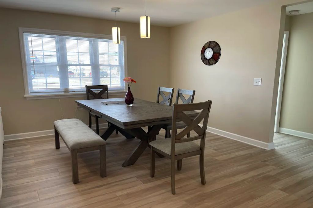 Un coin repas comprenant une table en bois, quatre chaises, un banc, des luminaires suspendus et une fenêtre avec des stores.