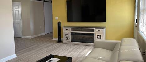 Een woonkamer met een beige bank, een flatscreen-tv boven een open haard, en een schuifdeur op de achtergrond.