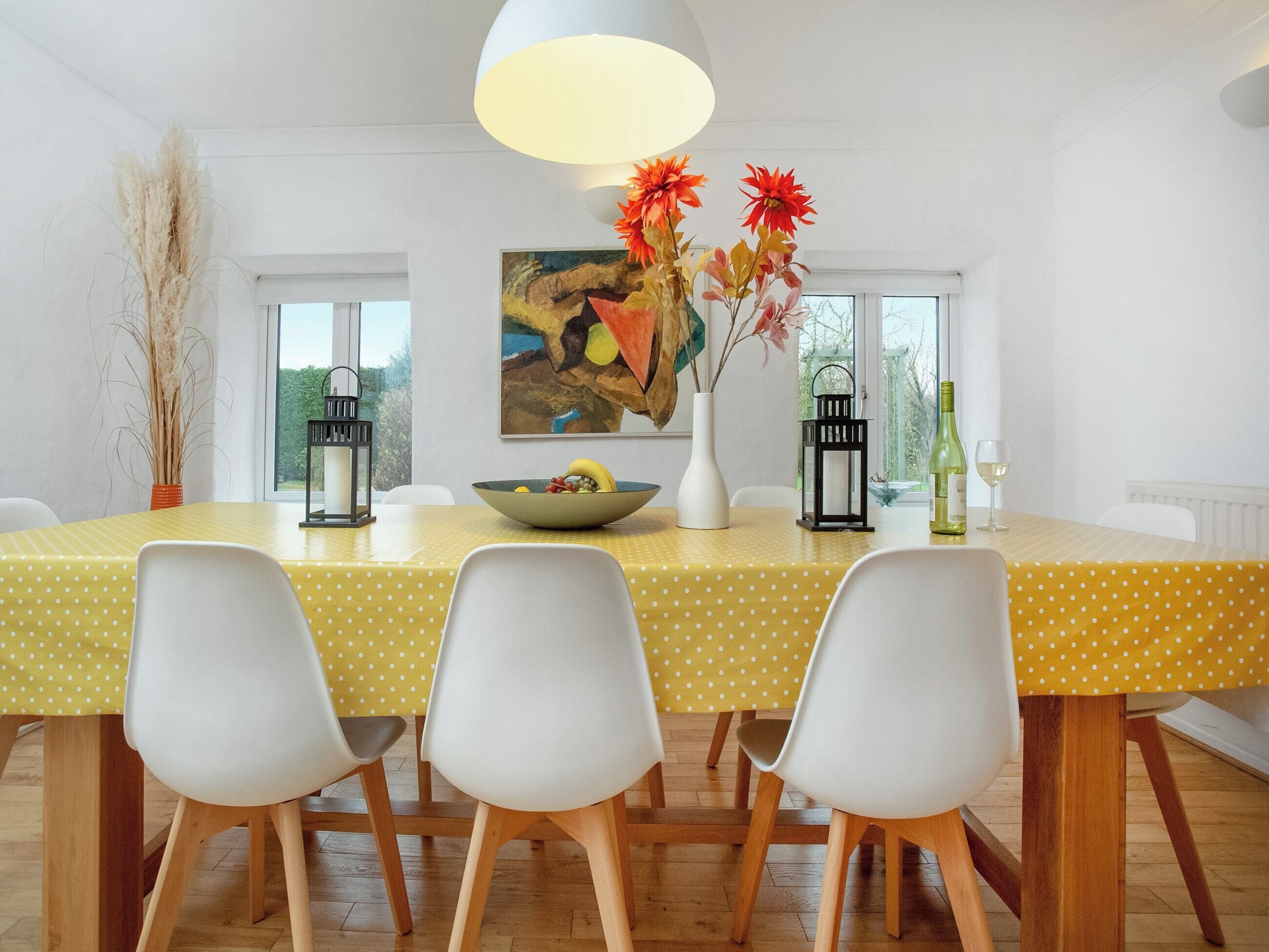 A dining area with a yellow table, white chairs, a vase with flowers, and a painting on the wall.