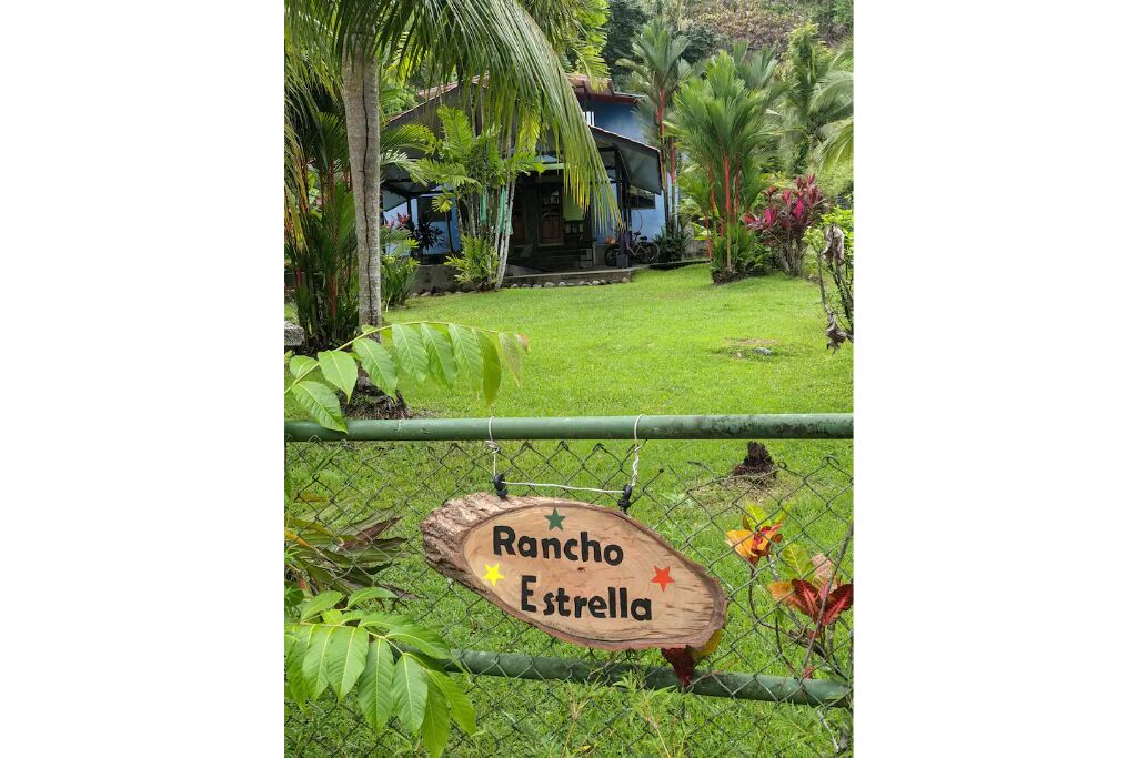 A wooden sign with "Rancho Estrella" written on it, attached to a chain-link fence with greenery around.