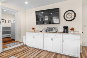A modern kitchen with white cabinetry, a flat-screen TV mounted above the counter, a wall clock, and a coffee maker. - Olver Farm Guest House : Park-Like Setting (Romulus)
