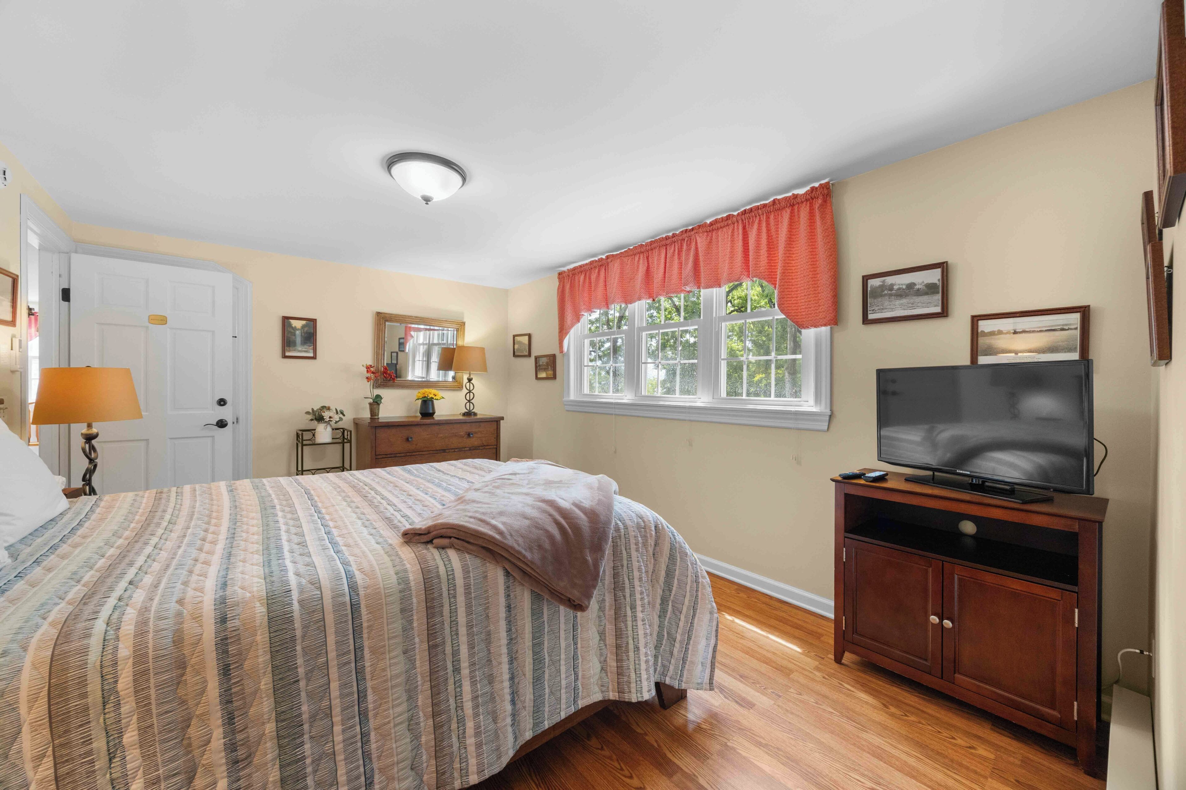 Une chambre à coucher avec un lit, une commode, un téléviseur et une fenêtre avec des rideaux.