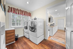 A laundry room with two front-loading washers and dryers, a wooden cabinet, a trash can, and a window with blinds.