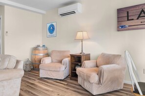 A living room with two beige armchairs, a wooden coffee table, a lamp, a wine barrel, and a wall-mounted air conditioner. - Olver Farm Guest House : Park-Like Setting (Romulus)