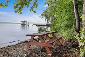 A picnic table on a pebbled shore with a dock and a house in the background.