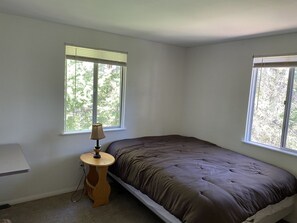 Quarto pequeno com uma cama de solteiro, janela em uma das paredes e uma luminária sobre uma mesinha.